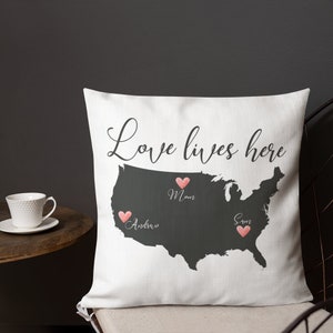 May include: White throw pillow with a dark gray map of the United States. The words "Love lives here" are written in cursive. Pink hearts mark locations with names "Mom", "Andrew", and "Sam".