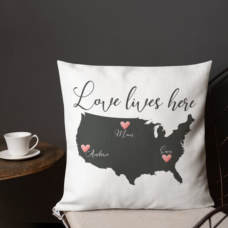 May include: White throw pillow with a dark gray map of the United States. The words "Love lives here" are written in cursive. Pink hearts mark locations with names "Mom", "Andrew", and "Sam".