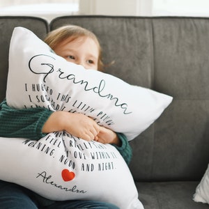 May include: A white pillow with black text that reads "Grandma" and "I hugged this little pillow" and "I squeezed it really tight" and "I feel you within it" and "morning, noon, and night." The pillow has a red heart and the name "Alexandra" printed on it. A young girl is holding the pillow and smiling.