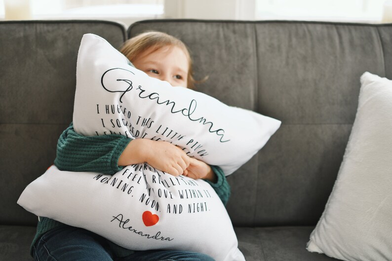 May include: A white pillow with black text that reads "Grandma" and "I hugged this little pillow" and "I squeezed it really tight" and "I feel you within it" and "morning, noon, and night." The pillow has a red heart and the name "Alexandra" printed on it. A young girl is holding the pillow and smiling.
