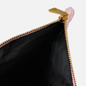 Puede incluir: Un neceser rosa parcialmente abierto con cremallera dorada. El interior es negro. La cremallera es dorada y la bolsa est&aacute; hecha de un material suave y texturizado. La bolsa est&aacute; vac&iacute;a y abierta.
