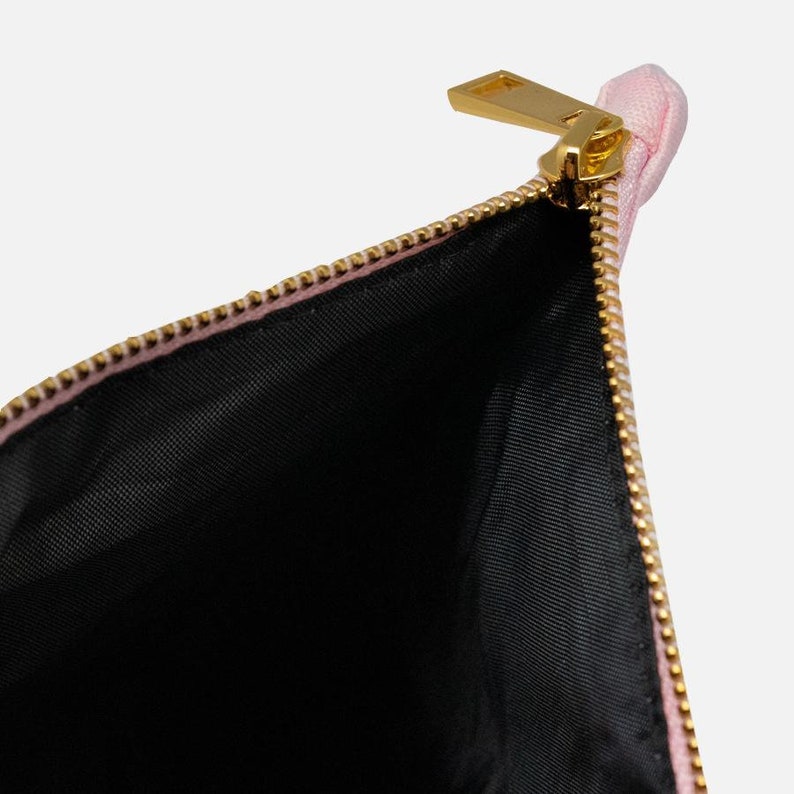 Puede incluir: Un neceser rosa parcialmente abierto con cremallera dorada. El interior es negro. La cremallera es dorada y la bolsa est&aacute; hecha de un material suave y texturizado. La bolsa est&aacute; vac&iacute;a y abierta.