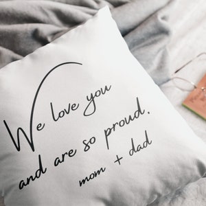 Pu&ograve; includere: Cuscino quadrato bianco con scritta nera "We love you and are so proud. mom + dad." Il cuscino &egrave; su un letto con una coperta grigia. Un libro e occhiali sullo sfondo.