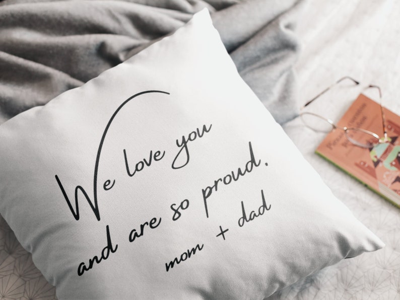 Pu&ograve; includere: Cuscino quadrato bianco con scritta nera "We love you and are so proud. mom + dad." Il cuscino &egrave; su un letto con una coperta grigia. Un libro e occhiali sullo sfondo.