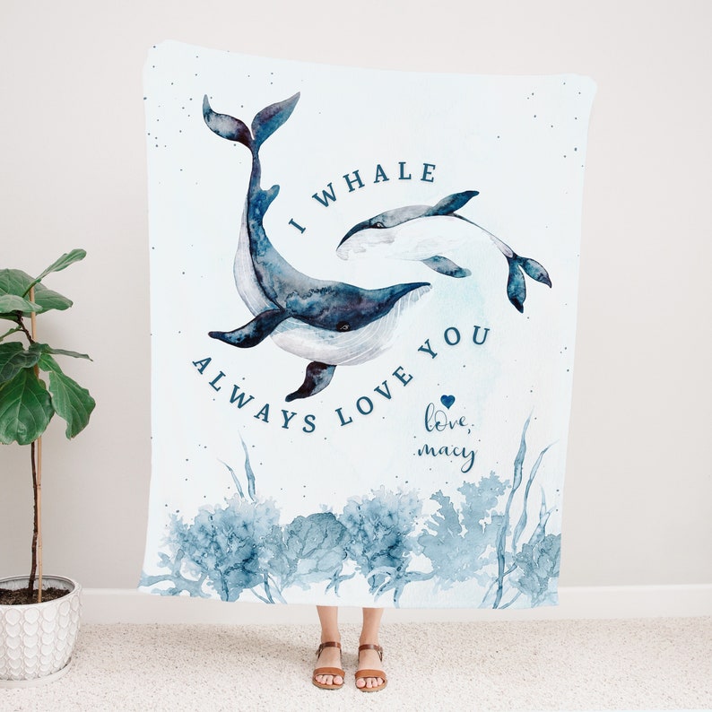 May include: A white blanket featuring a watercolor design of two whales in shades of blue. The blanket has the text "I whale always love you" and "love, macy" in blue script. The design includes blue watercolor coral and seaweed.