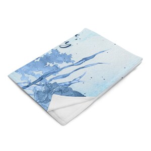 May include: A soft, folded throw blanket featuring a watercolor design in shades of blue. The artwork depicts abstract floral and plant-like shapes with the word "Joy" in cursive. The blanket has a white underside.