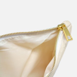 Puede incluir: Primer plano de una bolsa de lona blanquecina abierta, con cremallera y tirador dorados. El forro interior es de color beige claro. La cremallera est&aacute; completamente abierta, revelando el interior.
