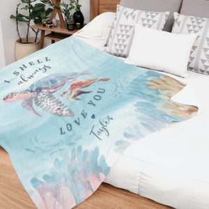 May include: A light blue blanket with a watercolor design featuring sea turtles and coral. The blanket has the text "I shell always love you Taylor" in a script font. The blanket is folded on a bed with white sheets and pillows.