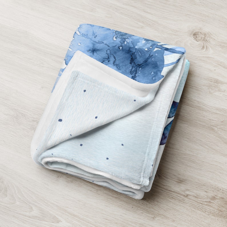 May include: A folded, soft blanket with a watercolor design in shades of blue. The blanket features a light blue base with darker blue watercolor splotches and small navy blue dots. The blanket is folded neatly, showcasing the design.