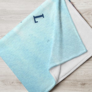 May include: A folded throw blanket with a light blue watercolor design. The blanket has a white underside and a blue letter "L" in the corner. The blanket is folded on a light wood surface.