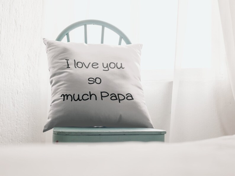 Pu&ograve; includere: Un cuscino quadrato bianco con la scritta "I love you so much Papa" in corsivo nero. Il cuscino &egrave; su una sedia azzurra, vicino a una finestra luminosa.