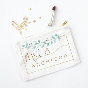 Puede incluir: Bolsa de cosm&eacute;ticos de lona blanca con cremallera dorada, con un dise&ntilde;o floral en acuarela y el texto "Mrs Anderson" en dorado. Tambi&eacute;n hay un labial y confeti dorado.