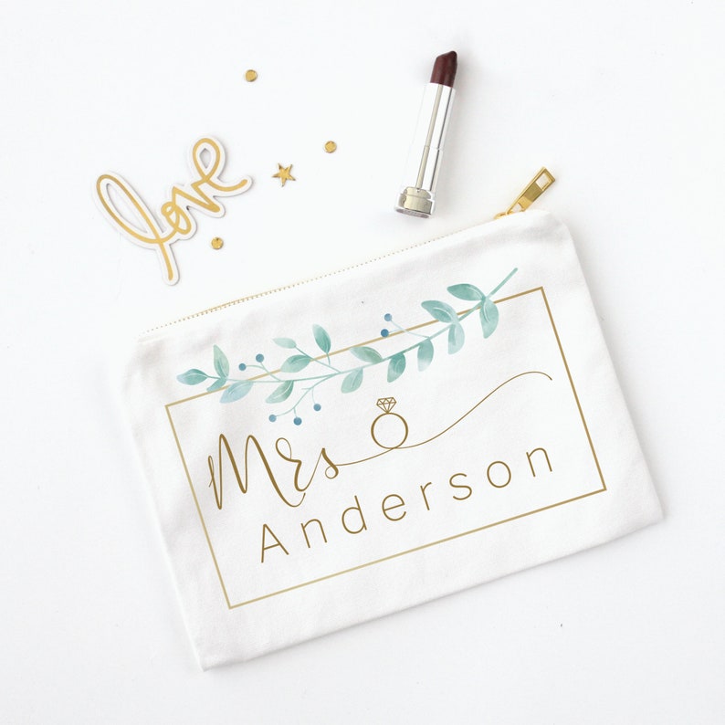 Puede incluir: Bolsa de cosm&eacute;ticos de lona blanca con cremallera dorada, con un dise&ntilde;o floral en acuarela y el texto "Mrs Anderson" en dorado. Tambi&eacute;n hay un labial y confeti dorado.
