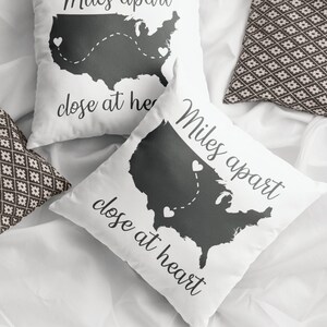 May include: Two white throw pillows with a black outline of the United States map and the text "Miles apart, close at heart." The pillows have a dotted line with hearts connecting different locations on the map. Two patterned pillows are also visible.
