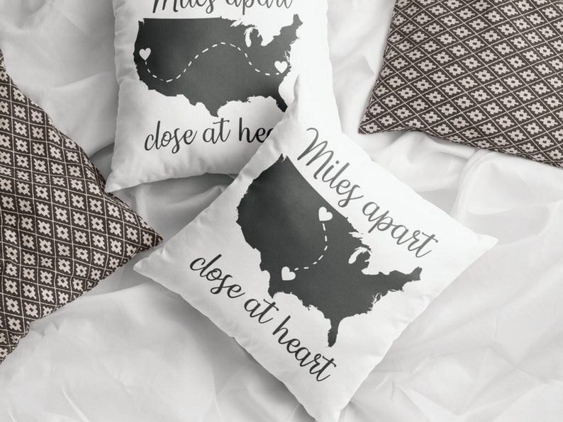 May include: Two white throw pillows with a black outline of the United States map and the text "Miles apart, close at heart." The pillows have a dotted line with hearts connecting different locations on the map. Two patterned pillows are also visible.