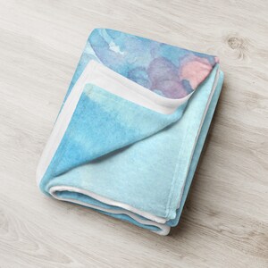 May include: Folded throw blanket with a watercolor design in shades of blue, purple, and pink. The blanket has a soft, plush texture and a white border. The blanket is folded neatly, showcasing the colorful pattern.