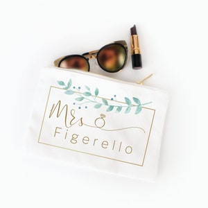 Puede incluir: Un neceser de lona blanca con cremallera dorada, las palabras "Mrs. Figarello" en escritura dorada y un dise&ntilde;o floral en acuarela. Gafas de sol y l&aacute;piz labial colocados encima de la bolsa.