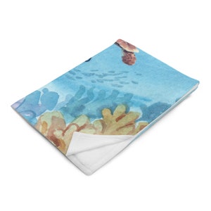 May include: A soft, folded blanket featuring a watercolor design of an underwater scene. The design includes a sea turtle, coral, and fish in shades of blue, tan, and white. The blanket has a white underside.
