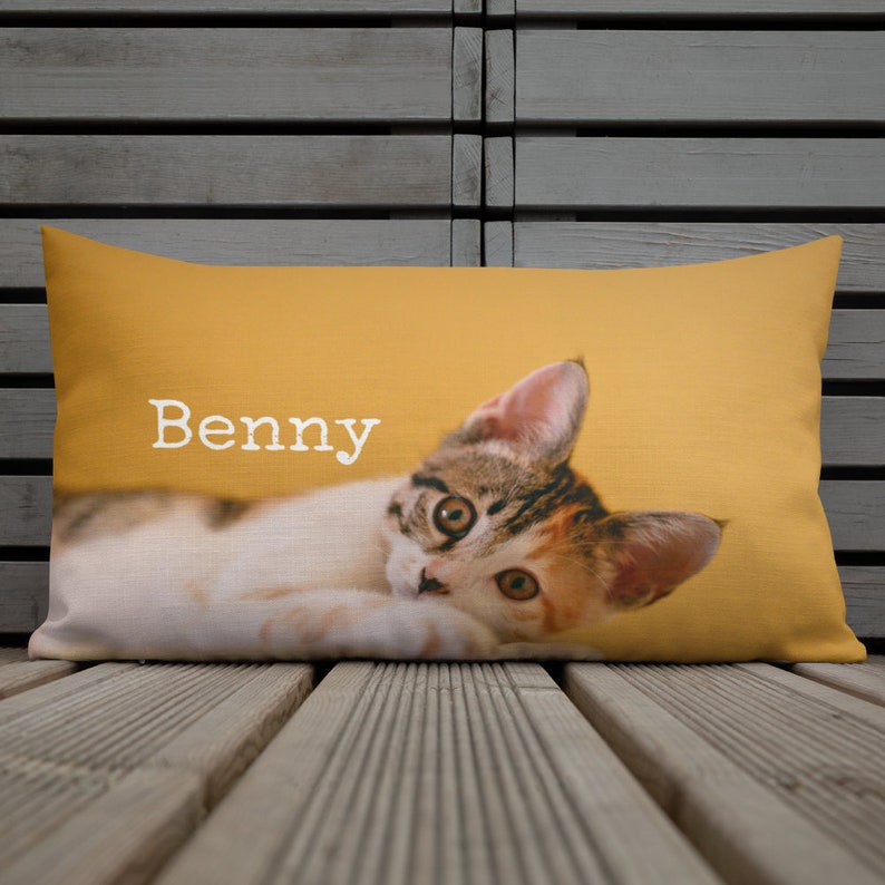 May include: Rectangular pillow with a photo of a calico cat named Benny on a mustard yellow background. The cat is lying down, looking at the viewer. The pillow is on a wooden surface.