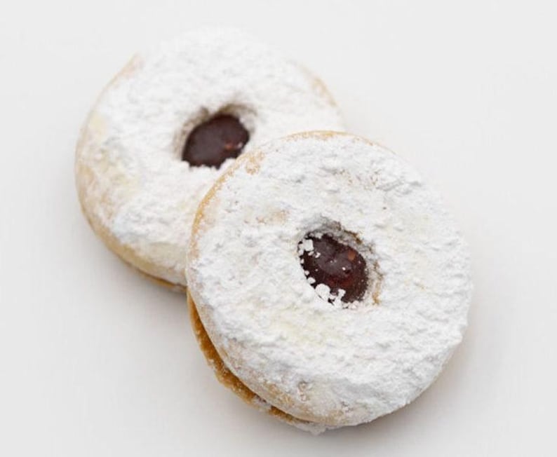 Op de afbeelding: Twee ronde koekjes, bedekt met poedersuiker, met een donkere vulling in het midden. De koekjes hebben een lichtbruine kleur en zijn royaal bestrooid met witte poedersuiker, een zoete traktatie.
