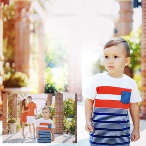 May include: A young boy wearing a white, red, and blue striped shirt with a denim pocket. He is standing in front of a brick wall with green plants in the background.