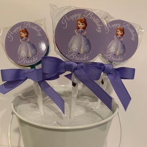 May include: Purple and white birthday lollipops with a princess character and the text "Happy Birthday Brooke" on each lollipop. The lollipops are arranged in a white bucket with purple ribbon bows.