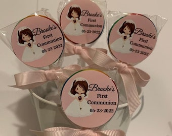 May include: Four pink and white lollipops with a girl in a white dress and a cross on a pink background. The lollipops have a white ribbon tied around the stick. The text on the lollipops reads "Brooke's First Communion 05-23-2022".