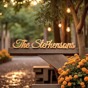 May include: Wooden sign with the text "The Stephensons" in a cursive font, placed on a wooden structure. The sign is in a natural wood color. The background features a blurred outdoor scene with string lights and orange flowers.