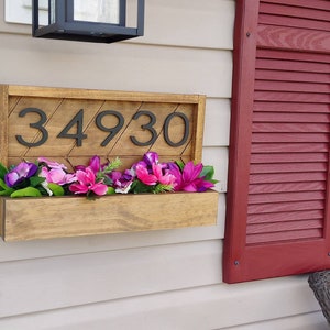 May include: A wooden house number sign with the number 34930 in black lettering. The sign is mounted on a white wall and has a planter box underneath filled with pink and purple flowers.