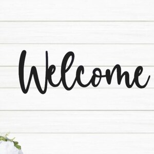May include: Black metal welcome sign with a cursive font on a white wood background.