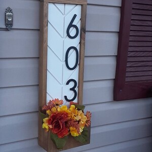May include: A wooden house number sign with a white chevron pattern and black numbers "603". The sign has a planter box with fall-colored artificial flowers.