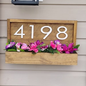 May include: Wooden address sign with the numbers "4198" in white. The sign has a brown wooden frame with a geometric pattern and a built-in planter box filled with pink and purple flowers.