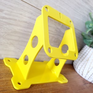 May include: A bright yellow metal bracket with multiple holes and a rectangular cutout. The bracket is designed for mounting and has a sturdy construction.