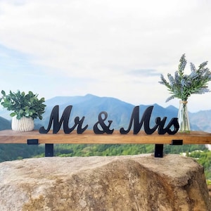 May include: A wooden shelf with black metal brackets, displaying a black "Mr. & Mrs." sign. The shelf is placed on a large rock with a blurred background of mountains and sky.