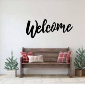 May include: A black metal "Welcome" sign hangs on a white wall above a wooden bench with red and white plaid throw pillows. A small green Christmas tree sits on either side of the bench.