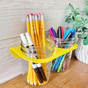May include: Two glass jars with yellow lids are filled with pencils and pens. The jars are sitting on a wooden surface. A potted plant is in the background.