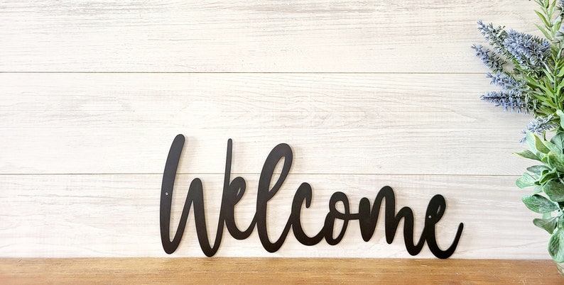 May include: Black metal welcome sign with a cursive font. The sign is mounted on a white wood background.