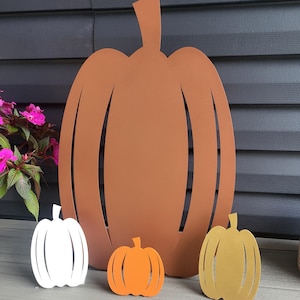 May include: A large brown metal pumpkin cutout stands behind three smaller pumpkin cutouts. The smaller ones are white, orange, and gold. Each pumpkin features a flat, stylized design with vertical cutouts representing the segments of the fruit.