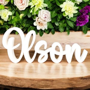 May include: A white wooden sign that spells out the name "Olson" in a cursive font. The sign is displayed on a wooden table, with a backdrop of colorful flowers.
