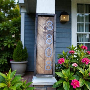 May include: Wooden address sign with the numbers 3808 in silver. The sign has a dark wooden frame and a chevron pattern on the wood. The sign is mounted on a white pillar, with pink flowers and green plants in the foreground.