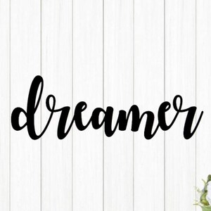 May include: Black cursive word "dreamer" on a white wood background.