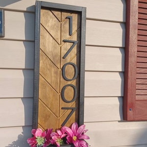 May include: A wooden house number sign with black trim and a chevron pattern. The number 1707 is displayed in black lettering. A black planter box with pink flowers is attached to the bottom of the sign.