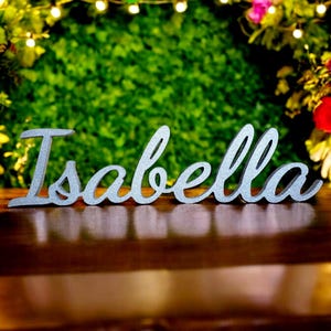 May include: Silver glitter name sign with the name "Isabella" spelled out in cursive letters. The sign is on a wooden surface with a green and floral background.