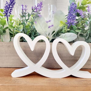 May include: A white, intertwined heart-shaped decorative object. The sculpture is placed in front of a wooden box filled with greenery and purple flowers. The hearts are smooth and have a modern design, perfect for home decor.