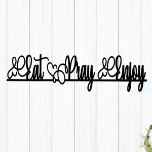 May include: Black metal wall art with the words "Eat Pray Enjoy" in a cursive font. The art piece is mounted on a white wooden background. The letters are connected and have decorative swirls.