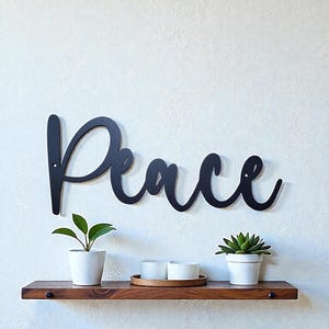 May include: Black metal wall art spelling the word "Peace" in a cursive font. The sign is mounted on a white wall above a wooden shelf. The shelf holds two small potted plants, candles, and a wooden tray.