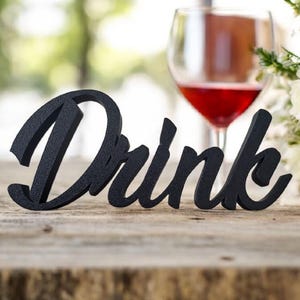 May include: Black wooden sign with the word "Drink" in a cursive font. The sign is sitting on a wooden table with a glass of red wine and a bouquet of white flowers in the background.