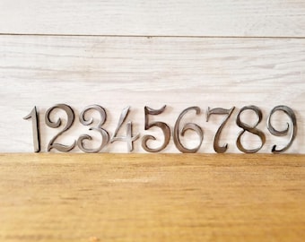 May include: A set of nine silver metal numbers, 1 through 9, with a decorative, cursive font. The numbers are arranged in a row on a wooden surface.