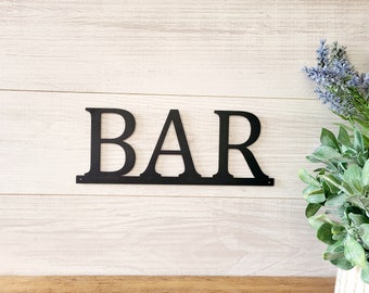 May include: Black metal sign with the word "BAR" in all caps. The sign is mounted on a white wood wall.