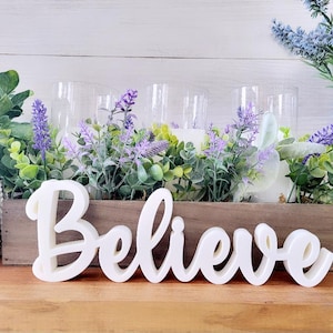 May include: White wooden sign with the word "Believe" in cursive lettering. The sign is sitting on a wooden table in front of a centerpiece of greenery and lavender.